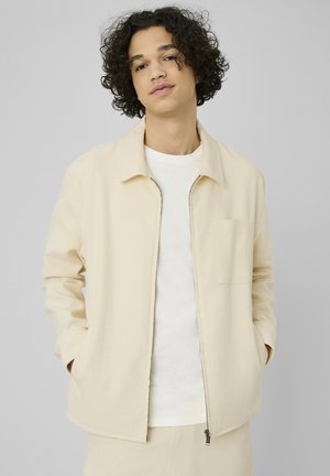 Jeune homme aux cheveux bouclés portant une veste zippée écrue sur une chemise blanche, debout devant un fond gris clair.