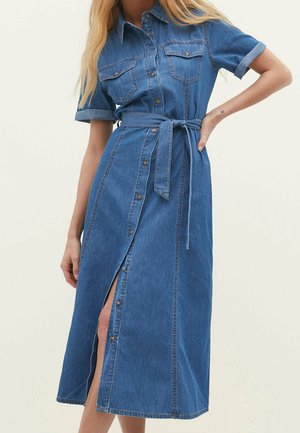 Vrouw draagt een mid-lange blauwe spijkerjurk met korte mouwen, een borstzakken en een tailleceintuur, staat met de hand op de heup.