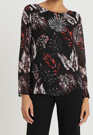 Kvinde iført en sort bluse med røde, hvide og grå tropiske bladmotiver og lange gennemsigtige ærmer, kombineret med sorte bukser.