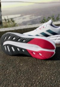 Des chaussures de sport avec une tige blanche ornée de rayures cyan, une semelle texturée noire et rouge, et des trous de ventilation pour une meilleure respirabilité.
