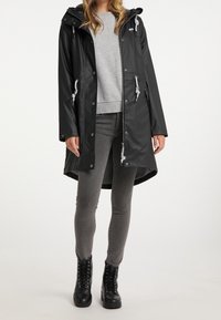 Imperméable noir avec capuche, doté de cordons de serrage, d'une fermeture éclair frontale complète et de deux poches latérales, associé à un jean gris et des bottes noires.