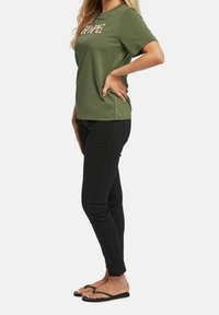 Woman standing in side view, wearing an olive green T-shirt with "HOPE" text, black fitted pants, and black flip-flops on a white background.