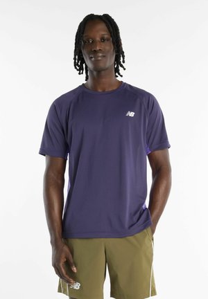 Man met gevlochten haar, die een paarse New Balance T-shirt en olijfgroene shorts draagt, staat tegen een effen witte achtergrond.