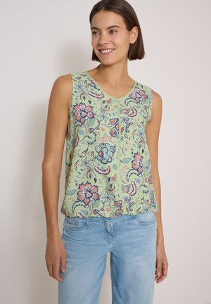 Frau trägt ein ärmelloses hellgrünes Oberteil mit bunten Blumenmustern und hellblaue Jeans, lächelnd und zur Seite schauend.