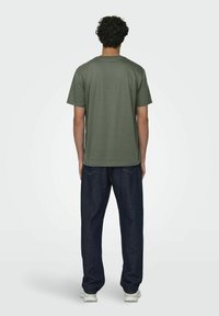 Camiseta de algodón verde con cuello redondo, mangas cortas y un corte relajado, combinada con pantalones de mezclilla azul oscuro y zapatillas blancas.