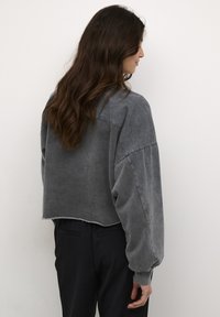 Apykaklė, per didelė pilka džemperis su ilgomis rankovėmis, išblukusi tekstūra ir brutali apdaila. Suderinta su juodomis kelnėmis, matoma iš užnugario.