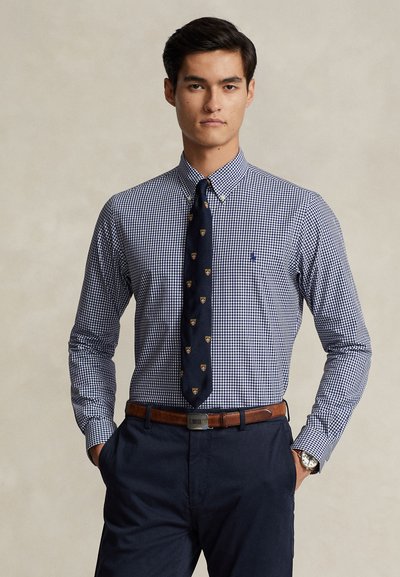 Junger Mann, der ein blau-weiß kariertes Hemd mit Knöpfen, eine marineblaue Krawatte mit kleinen Emblemen, einen braunen Gürtel, marineblaue Hose und eine Armbanduhr trägt.