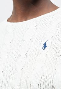 Gros plan d'une personne portant un pull en maille torsadée blanche avec un logo de joueur de polo brodé en bleu sur la poitrine.