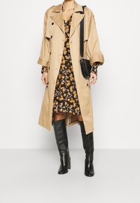Beige Trenchcoat über einem schwarzen Blumenkleid. Knielange schwarze Stiefel. Der Mantel hat große Knöpfe und eine taillierte Taille.
