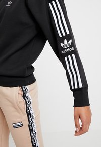 Svart sweatshirt med vit Adidas-logotyp och tre vertikala ränder på ärmarna. Matchas med beiga joggers med logotape på sidan.