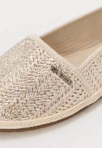 Flate slip-on sko i beige vevd stoff med metalliske detaljer, teksturert overflate og sydd kant. Har et merket metallsymbol.