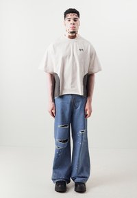 Hellbeige Oversized-T-Shirt mit grauen Akzenten, kombiniert mit zerrissenen blauen Weitbeinjeans mit Rissen und Farbspritzern. Schwarze klobige Schuhe.