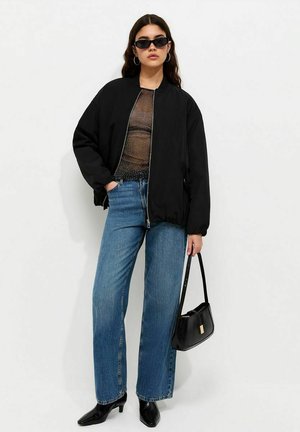 Mujer con chaqueta bomber negra, top negro transparente, vaqueros azules, zapatos negros de tacón, gafas de sol, pendientes de aro y sosteniendo un bolso negro.