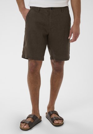 Mand iført mørkebrune knælange shorts og mørkebrune remsandaler med spænde, stående med den ene hånd i lommen.