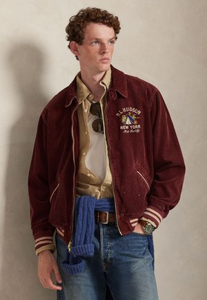 Bordeauxrød kordjakke med stribede manchetter, broderet logo, beige skjorte under, blå strikket trøje bundet om taljen og denimjeans.