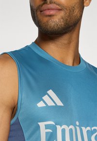 Débardeur de sport bleu avec un col rond. Affiche le logo "Adidas" en blanc avec trois bandes et l'impression "Emirates" à l'avant. Texture lisse.