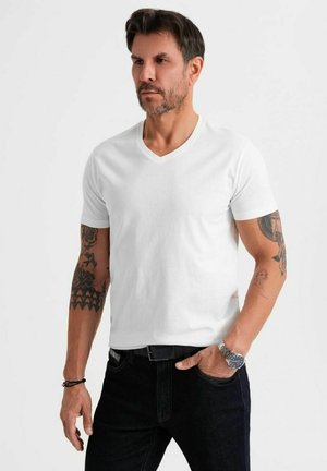T-shirt bianco in cotone a maniche corte con scollatura a V, dalla texture morbida, abbinato a jeans scuri e a un orologio d'argento, con tatuaggi visibili sulle braccia.