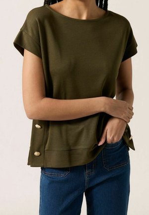 Kobieta w oliwkowej bluzce z krótkim rękawem, z złotymi guzikami po bokach i niebieskich dżinsach, z rękoma skrzyżowanymi w pasie.