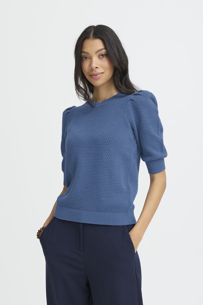 Femme aux cheveux longs et bruns portant un pull à manches courtes bleu texturé et un pantalon sombre, debout les mains dans les poches sur un fond blanc.