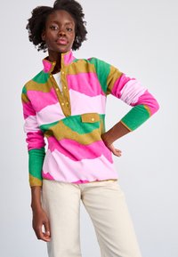 Pullover in pile colorato con motivi a onde in rosa, verde e oro. Presenta un colletto con zip a metà, dettagli con bottoni e una tasca frontale.