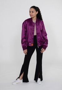 Paarse satijnen bomberjack met zijzakken, geribbelde manchetten en een rits. Gecombineerd met zwarte spijkerbroeken met split-zoom en witte sneakers.