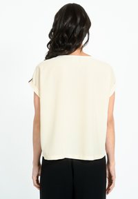 Īsām piedurknēm, apaļš kakla izgriezums topā gaiši smilškrāsā ar gludu tekstūru. Apvērsti manžeti un brīvs piegriezums. Valkāts virs melnām biksēm.