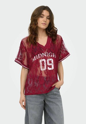 Jeune femme aux cheveux bruns ondulés portant un haut en dentelle bordeaux transparent avec le texte « MIDNIGHT 09 » et un jean bleu clair, main dans la poche.