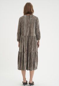 Kvinna som står med ryggen synlig, klädd i en lång, texturerad klänning i gråbeige med trekvartslånga ärmar och svarta klacksandaler.