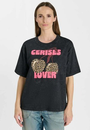 Donna che indossa una T-shirt nera con testo rosa "CERISES LOVER" e due ciliegie con stampa leopardata, abbinata a jeans azzurri chiari.