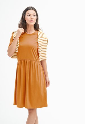 Braunes, knielanges Kleid mit runder Halslinie und geraffter Taille. Gestreifte, hellfarbige Jacke, die über die Schultern gelegt ist, sorgt für einen Kontrast.