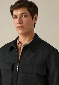 Black zip-up jacket with a pointed collar, two chest pockets, and a textured beige knit top underneath. Smooth fabric with minimal detailing.