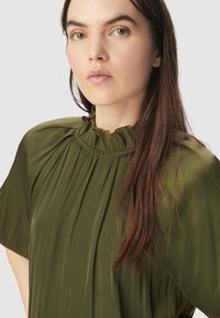 Blusa verde oliva con un diseño plisado, cuello alto con volantes y una tela suave y fluida de textura lisa.