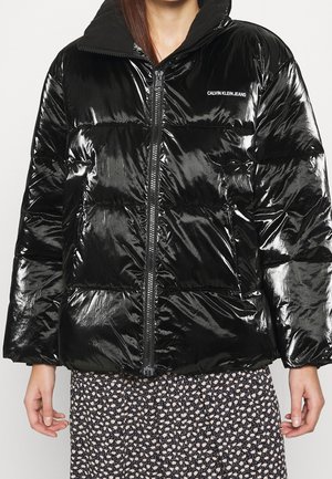 Femme portant une doudoune noire brillante avec une fermeture éclair à l'avant et le logo "Calvin Klein Jeans", associée à une jupe noire et blanche à motifs floraux.