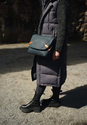 Teal messenger bag with orange accents, textured material, and an angular design, worn crossbody over a gray puffer vest and black pants.