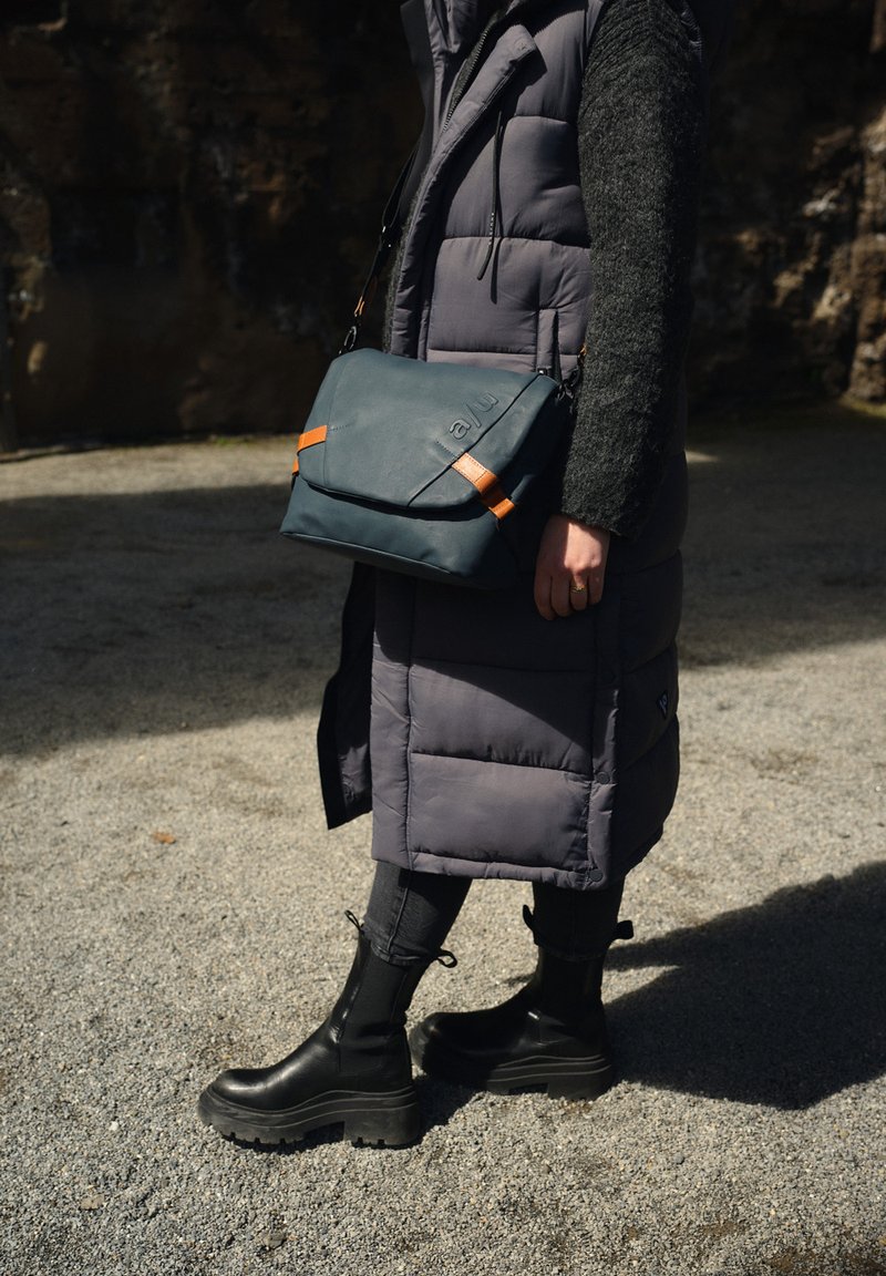 Teal messenger bag with orange accents, textured material, and an angular design, worn crossbody over a gray puffer vest and black pants.
