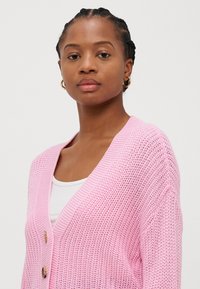 Adīts rozā kardigāns ar v-veida kaklu, divām lielām pogām un ribotām piedurknēm. Piemīt vaļīga, atvērta auduma struktūra.