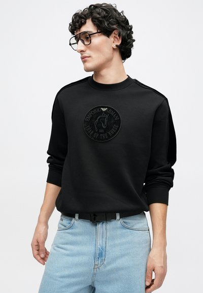 Melnā sporta džempera ar apaļu, reljefu emblēmas plāksteri ar zirga dizainu. Tam ir apaļa kakla izgriezums un stilīgas riboti aproces, valkātas kopā ar gaišām džinsa biksēm.