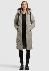 Lang puffet frakke i lys beige med chevron-quilting, stor hætte, sorte snore og sidelommer. Båret over sorte leggings og ankelstøvler.