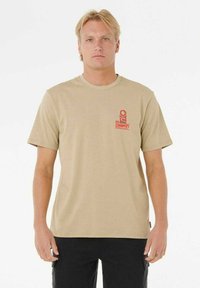 Camiseta de algodón beige con mangas cortas y cuello redondo. Presenta un diseño de logo rojo en la parte izquierda del pecho. Textura minimalista, corte recto.