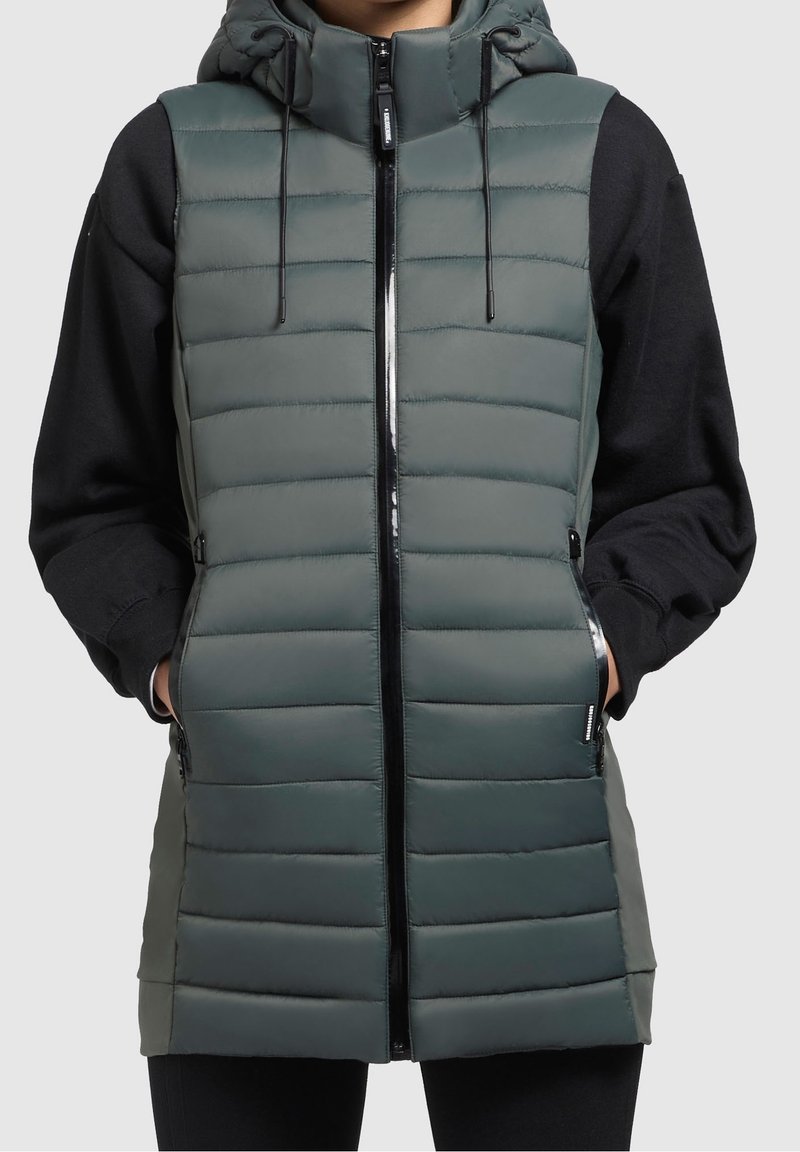Gilet imbottito verde scuro con zip frontale, cappuccio regolabile e tasche laterali. Presenta cuciture orizzontali e una finitura elegante.