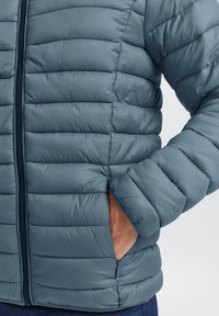 Blå isolert jakke med horisontalt quiltet design, glidelås og sidelommer. Glatt, lett materiale med en puffy tekstur.