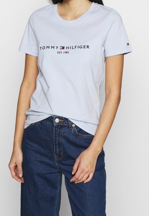 Mujer que lleva una camiseta azul claro de Tommy Hilfiger con logo y vaqueros azul oscuro, de pie con las manos relajadas a los lados.