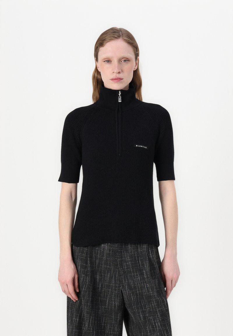 Top scurt cu maneci scurte, negru, cu guler înalt și închidere cu fermoar până la jumătate. Prezintă un logo minimal pe față, asortat cu pantaloni texturați.