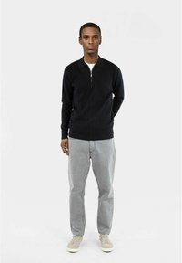 Felpa nera con zip e collo, abbinata a jeans grigi chiari e sneaker beige, creando un look casual e monocromatico.