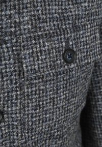 Wollen jas in een blauw, grijs en zwart ruitpatroon; voorzien van een borstzak en een zwarte knoopsluiting. Gestructureerde stof.