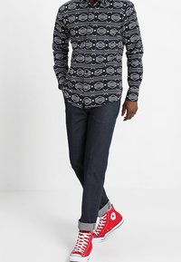 Marineblauw overhemd met witte geometrische patronen, gecombineerd met donkere spijkerbroek en rode hoge sneakers. Opgerolde mouwen van de broek.