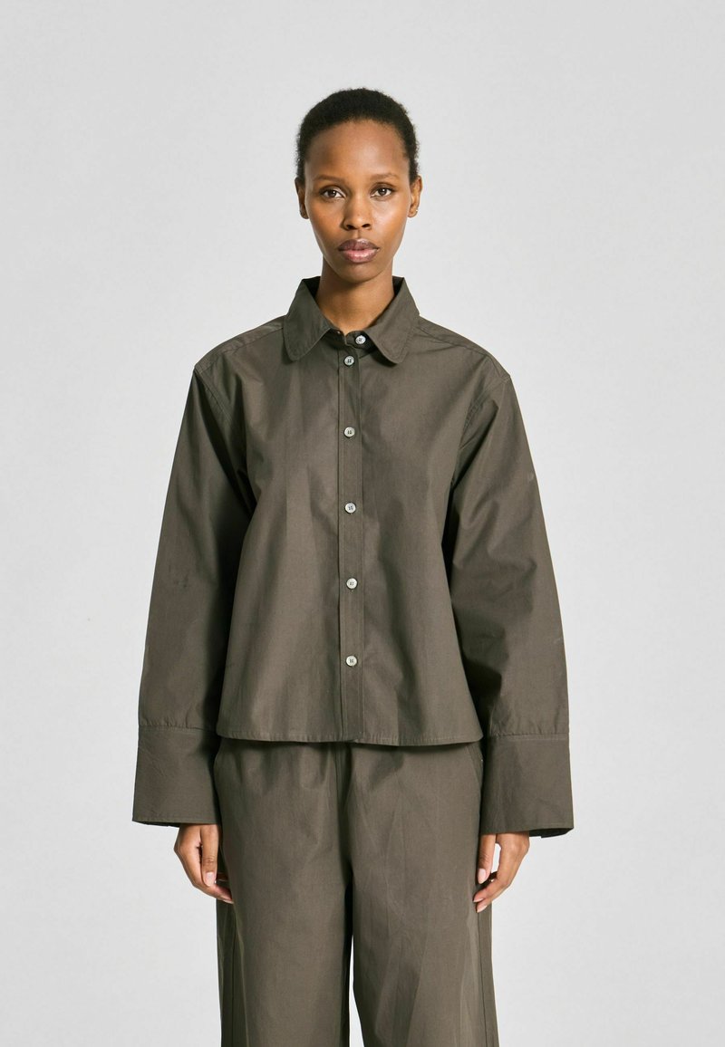 Femme debout face à l’avant, portant une chemise ample vert olive à boutons avec de larges poignets et un pantalon large assorti.