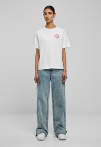 Hvit bomulls t-skjorte med korte ermer og et rosa grafisk trykk på venstre bryst. Kombinert med blå vide denimjeans og hvite sneakers.