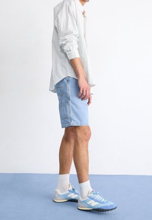 Homme portant un short en jean bleu clair, des chaussettes blanches, des baskets bleues et blanches et une chemise blanche à rayures manches longues, debout sur un sol bleu.