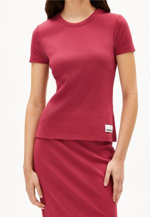 Kobieta ubrana w obcisły ciemnoróżowy prążkowany top z krótkim rękawem i dopasowaną spódnicę, stojąca z luźno opuszczonymi ramionami po bokach.